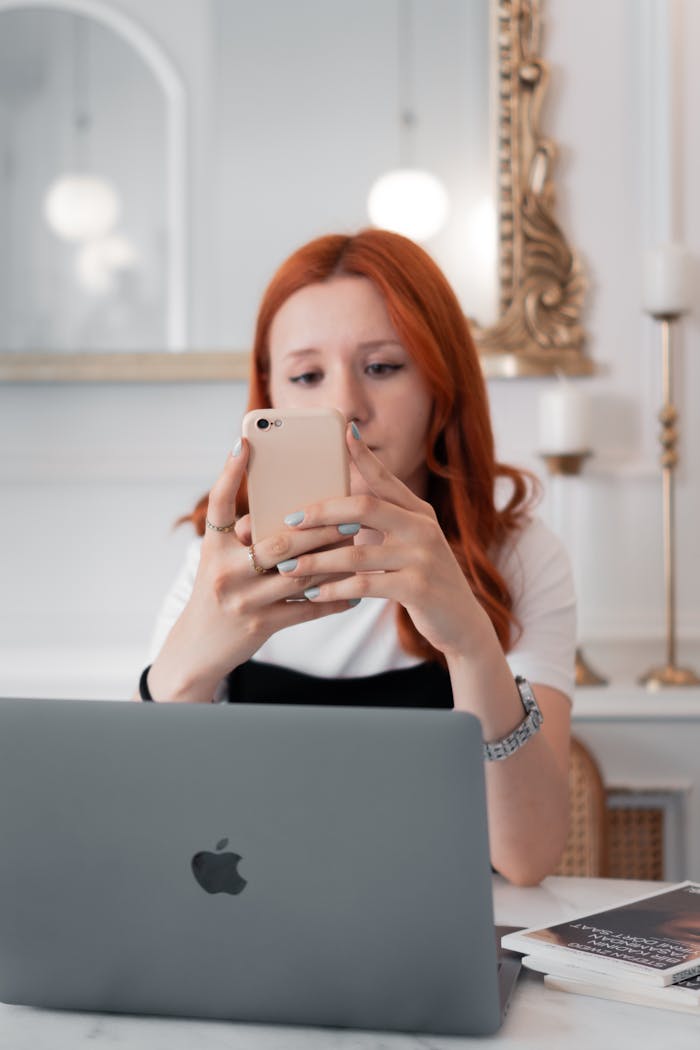 Young woman with red hair using a smartphone at a desk with a laptop in a stylish room.